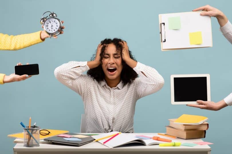 Jovem negra executiva, sentada na mesa de trabalhando cercada por mãos segurando telefone, relógio e outros acessórios de trabalhando, enquanto ela se mostra estressada, com a mão na cabeça.