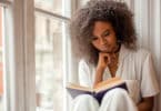 Uma garota negra lendo um livro no peitoril da janela.