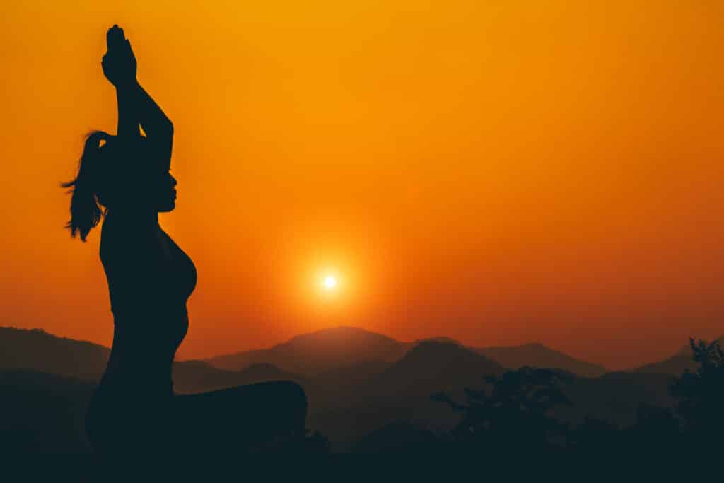 Mulher meditando em meio a natureza, com o pôr do sol ao fundo