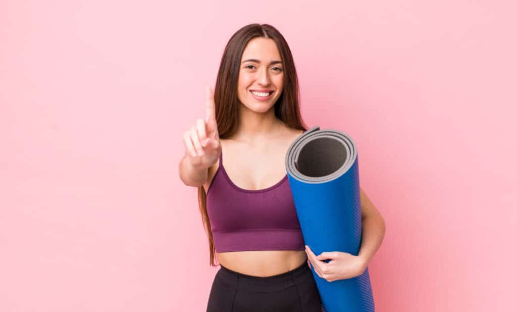 Mulher branca de cabelos longos e castanhos, sorrindo enquanto segura um tapete de yoga e faz o número 1 com uma das mãos.