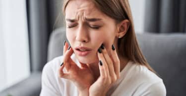 Jovem mulher com os olhos fechados e tocando as bochechas com as pontas dos dedos, como se estivesse com dor nos dentes.