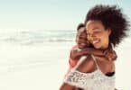 Mulher sorridente carregando uma garotinha, também sorridente, em suas costas. Elas estão em uma paisagem praiana.