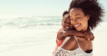 Mulher sorridente carregando uma garotinha, também sorridente, em suas costas. Elas estão em uma paisagem praiana.