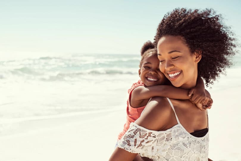Mulher sorridente carregando uma garotinha, também sorridente, em suas costas. Elas estão em uma paisagem praiana.