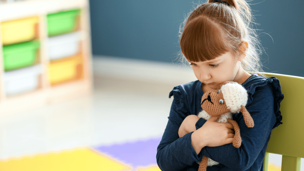 Menina ruiva segurando seu bichinho de pelúcia, sentada na mesa da escola com expressão de chateada.