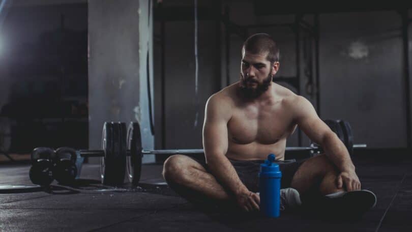 Imagem de um homem musculoso na academia sentado no chão e de cabeça baixa.