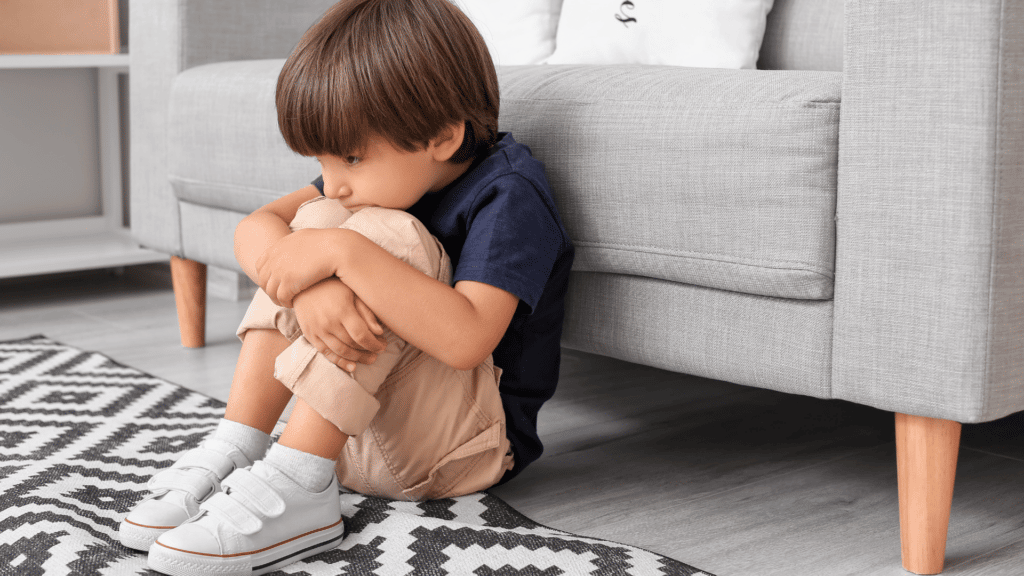 Menino branco sentado no chão a frente de um sofá, em posição fetal com expressão de desânimo.