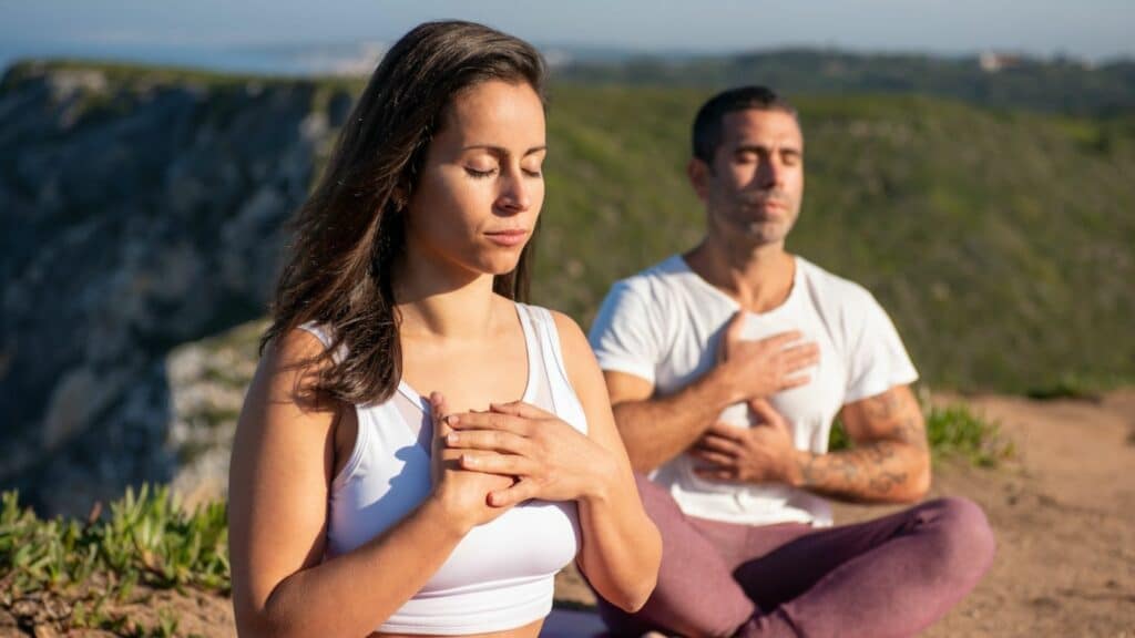 Imagem de um homem e uma mulher meditando em uma montanha, com os olhos fechados.