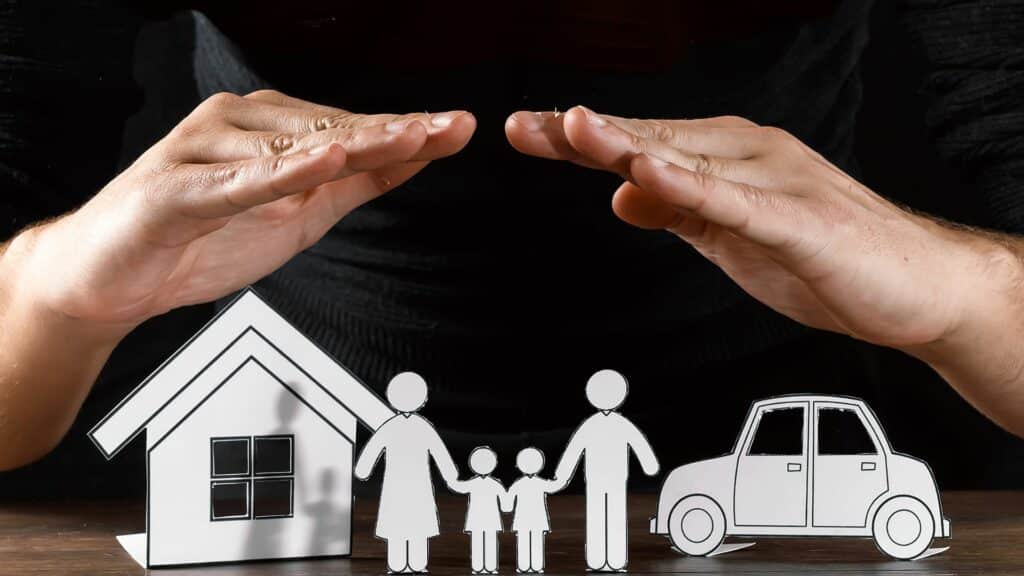 Imagem de duas mãos em cima de um recorte de papel representando a casa a família e um carro, as mãos protegem esse lar.