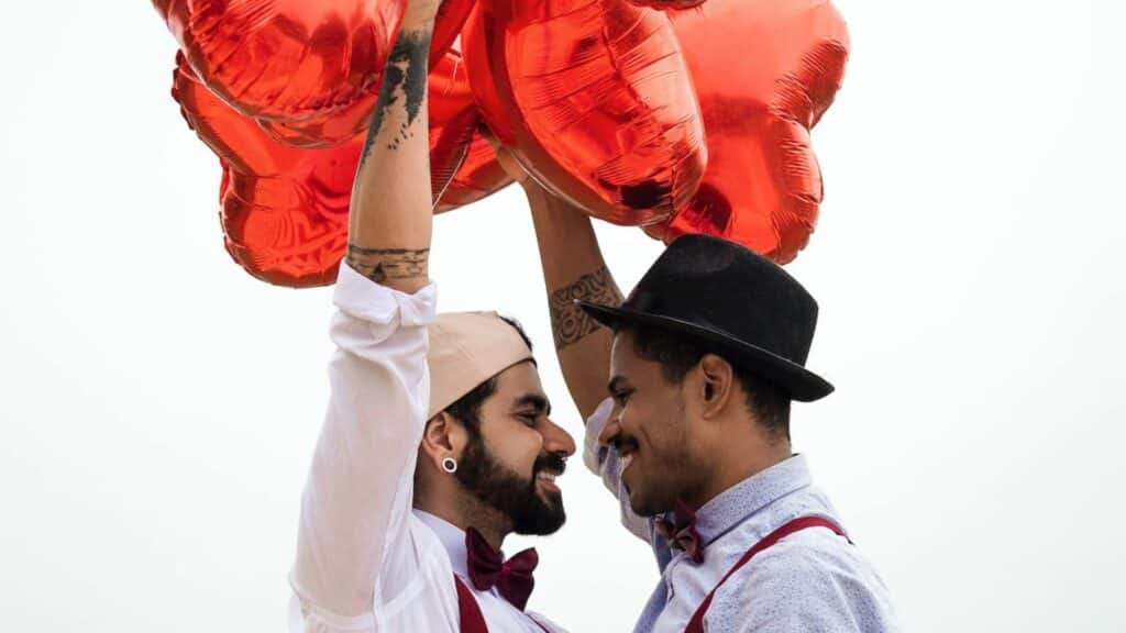 Imagem de um casal de homens se encarando e sorrindo e segurando 
balões em formato de coração acima de suas cabeças.
