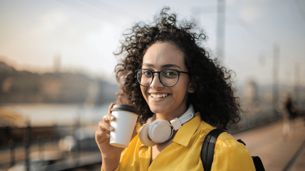 Mulher com fone de ouvido branco no pescoço, com um copo de café na mão sorrindo.