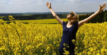 mulher com os braços erguidos e sorrindo em um campo cheio de flores amarelas.