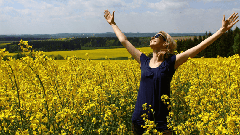 mulher com os braços erguidos e sorrindo em um campo cheio de flores amarelas.