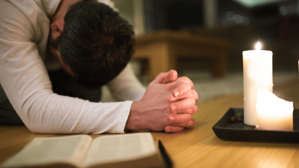 Homem de joelhos orando com vela e bíblia ao lado