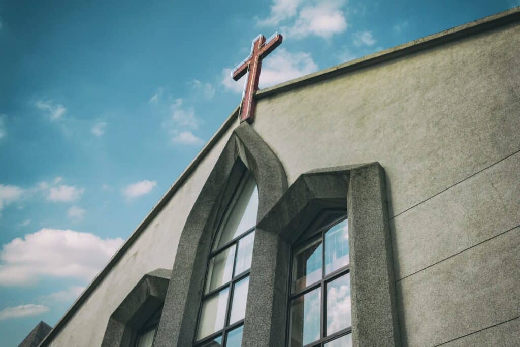 Fachada superior de uma igreja. São mostrada longas janelas e uma cruz marrom está posta entre elas e o telhado da construção.