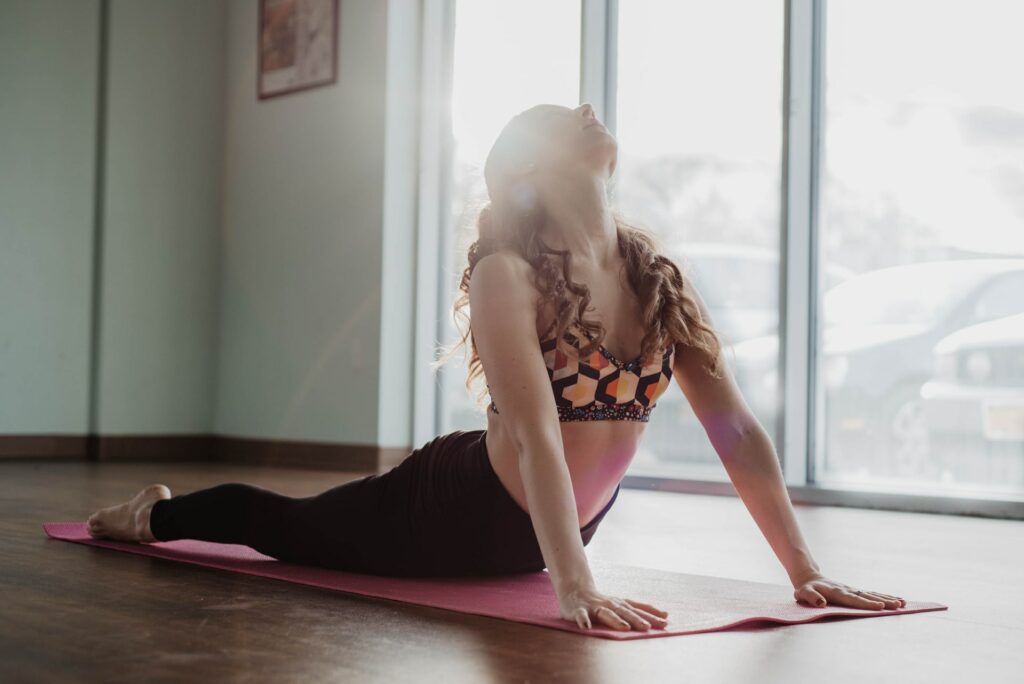 Mulher praticando a postura Urdhva Mukha Svanasana sobre seu tapete de yoga, em uma sala ampla e vazia.