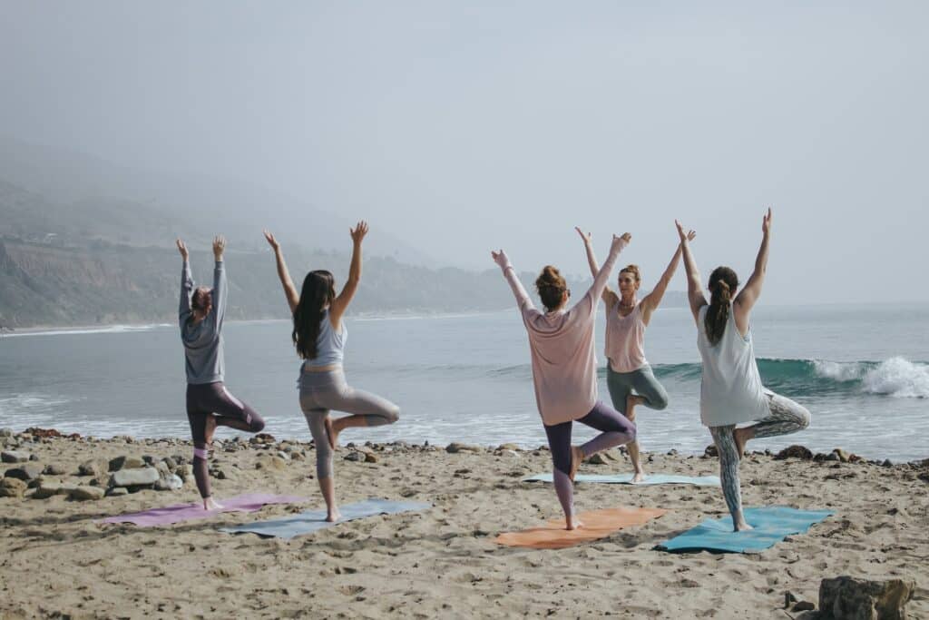 Grupo de quatro mulheres praticando yoga na praia, com uma instrutora.