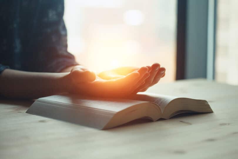 Mãos abertas voltadas para cima com uma luz amarela dentro dela, enquanto a mesma está apoiada sob a Bíblia.