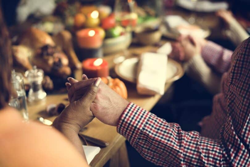 Recorte de uma família sentada à mesa com as mãos dada em momento de oração.