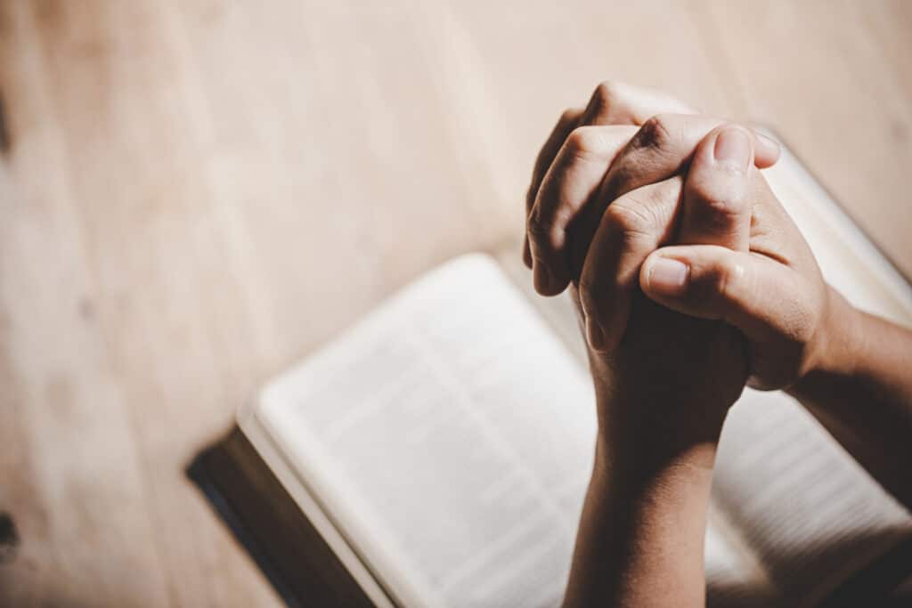 Mãos fazendo gesto de oração e ao fundo a Bíblia.