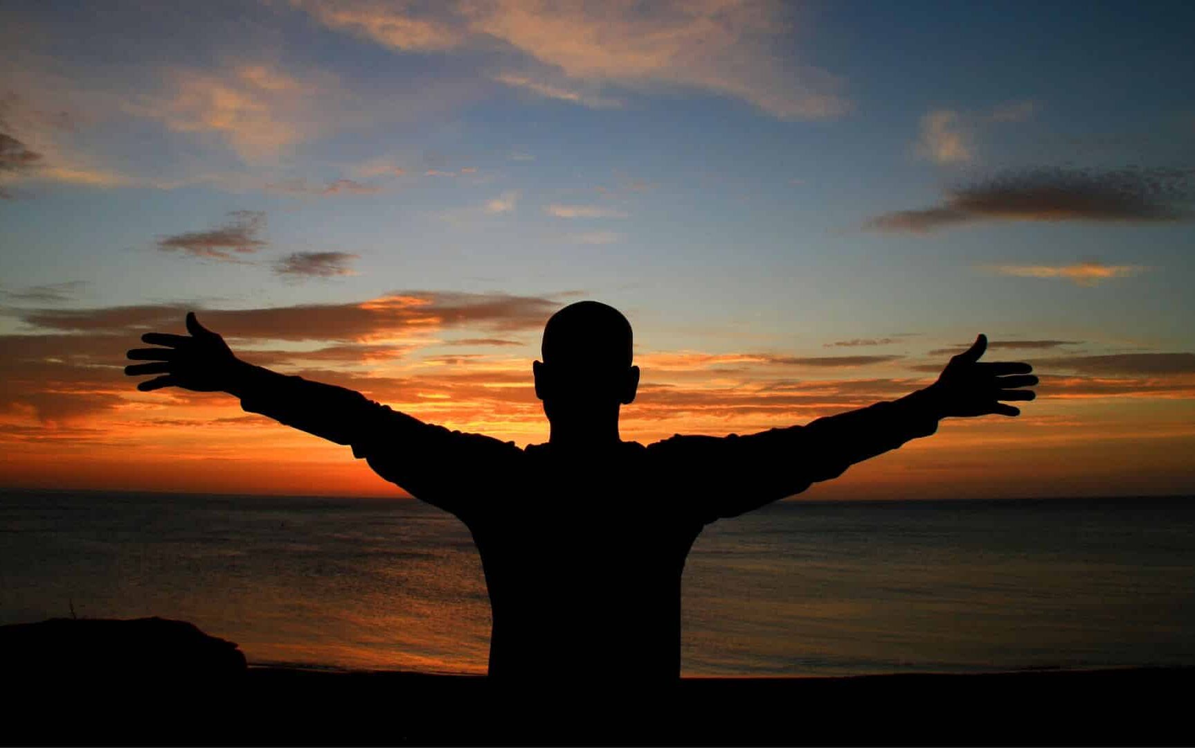 Um homem de braços erguidos à beira de uma montanha. Ao fundo, o pôr do sol.