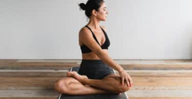 Mulher sentada sobre o tapete de yoga, em um chão de madeira, com suas pernas cruzadas e o tronco levemente torcido para sua esquerda.