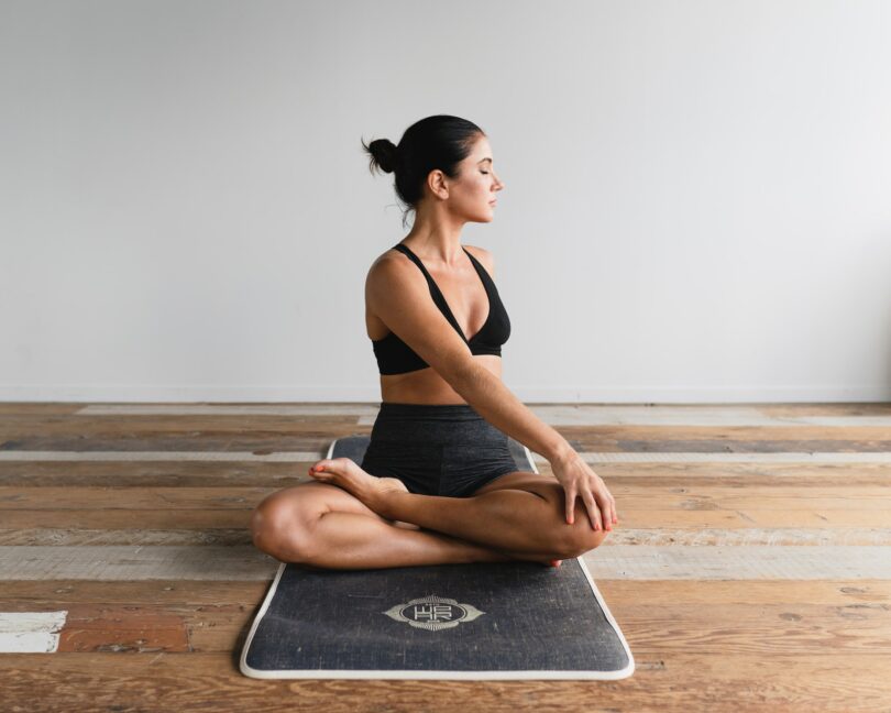 Mulher sentada sobre o tapete de yoga, em um chão de madeira, com suas pernas cruzadas e o tronco levemente torcido para sua esquerda.