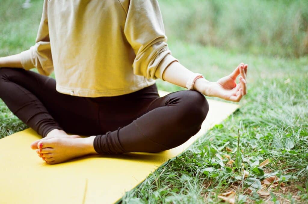 Mulher sentada em um tapete de yoga, em posição de lótus. O tapete está sobre um gramado e a foto mostra apenas de seu tórax para baixo.