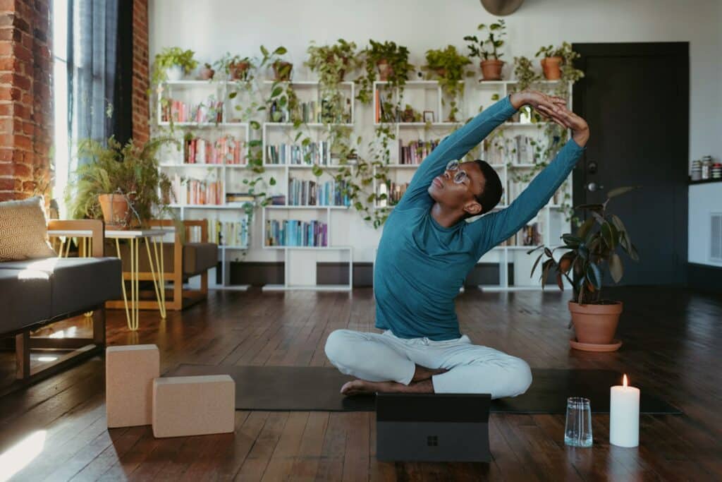 Homem sentado sobre um tapete de yoga, em meio à sala de sua casa. Ele está se alongando com os braços para o alto. À sua frente, um notebook e uma vela acesa.