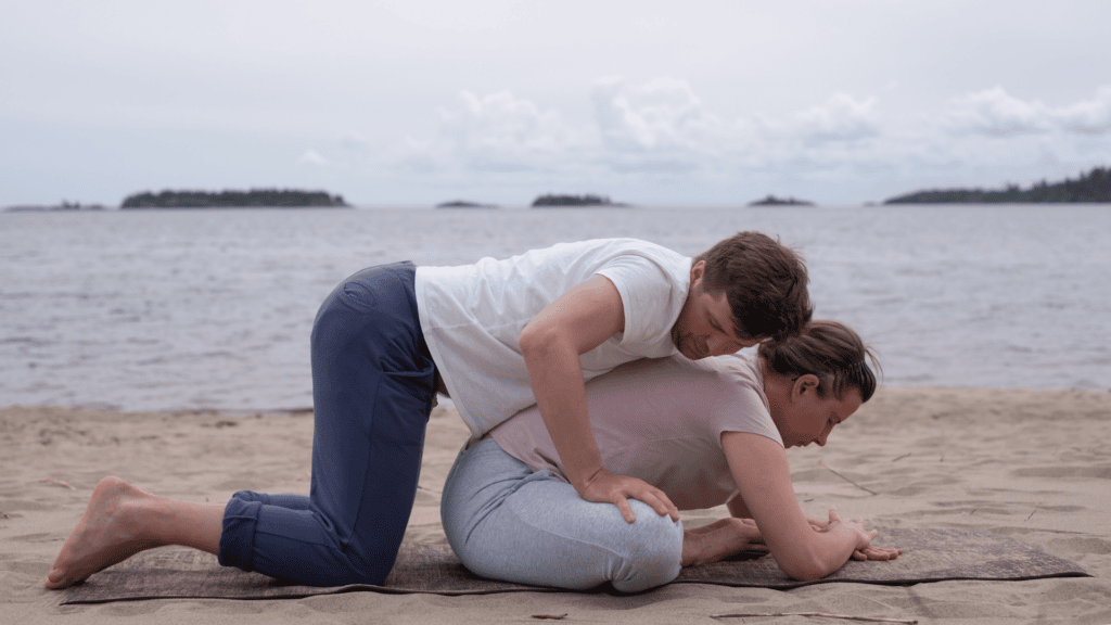 Um casal praticando yoga, enquanto a moça se encontra na postura da borboleta, o homem se apoia nas costas dela, afim de ajudar ela a alongar mais a coluna.