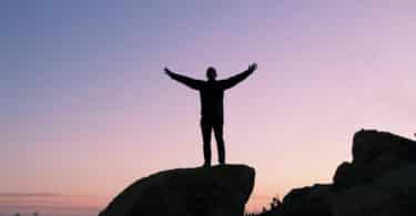 Imagem de um homem de braços abertos em cima de uma pedra e mostrando o céu arroxeado ao fundo
