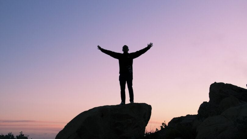 Imagem de um homem de braços abertos em cima de uma pedra e mostrando o céu arroxeado ao fundo