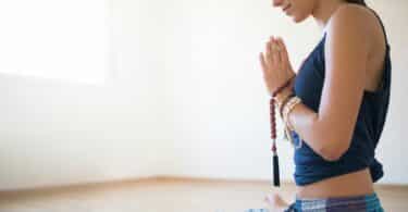 Mulher sentada em posição de lótus e com as mãos juntas em oração, de perfil.