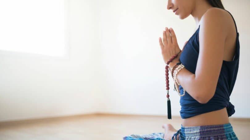 Mulher sentada em posição de lótus e com as mãos juntas em oração, de perfil.