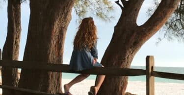 Imagem de uma moça sentada de costas em um cercado de frente para uma praia, do lado das árvores