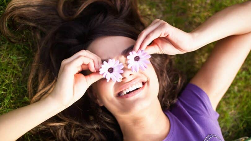 Imagem de uma mulher deitada no gramado, segurando duas flores na frente dos olhos