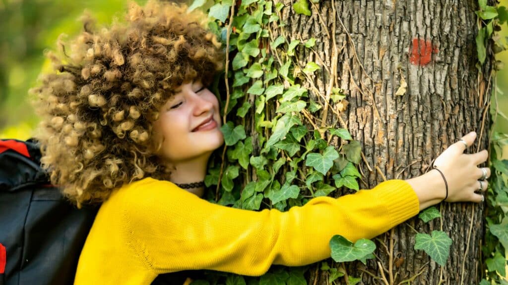 Imagem de uma mulher abraçando uma árvore e sorrindo de olhos fechados.