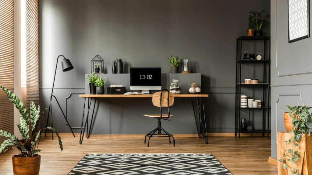 Imagem de um escritório com a parede cinza, uma mesa e cadeira de madeira e um computador em cima da mesa. Tem plantas espalhadas pela sala, uma luminária preta e uma estante de ferro na lateral perto da parede.