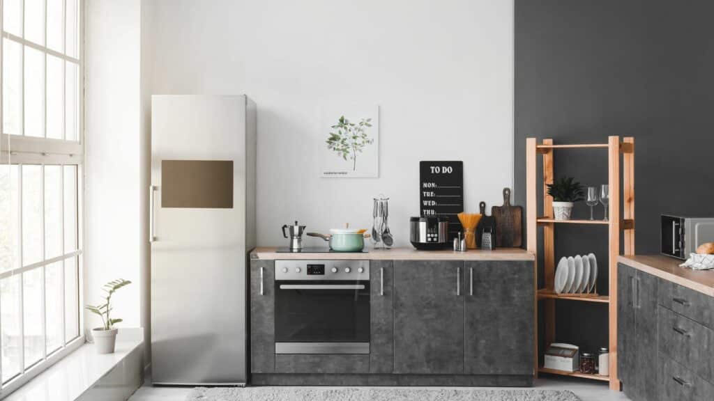Imagem de uma cozinha de cor clara e cinza, a geladeira está do lado da parede, logo ao seu lado o forno embutido em um armário de cor cinza escuro e outras peças de cozinha como pratos em uma estante. A janela da lateral é grande e clareia ainda mais o ambiente