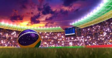 Imagem de um estádio de futebol, cheio e iluminado, e uma bola do Brasil no gramado.