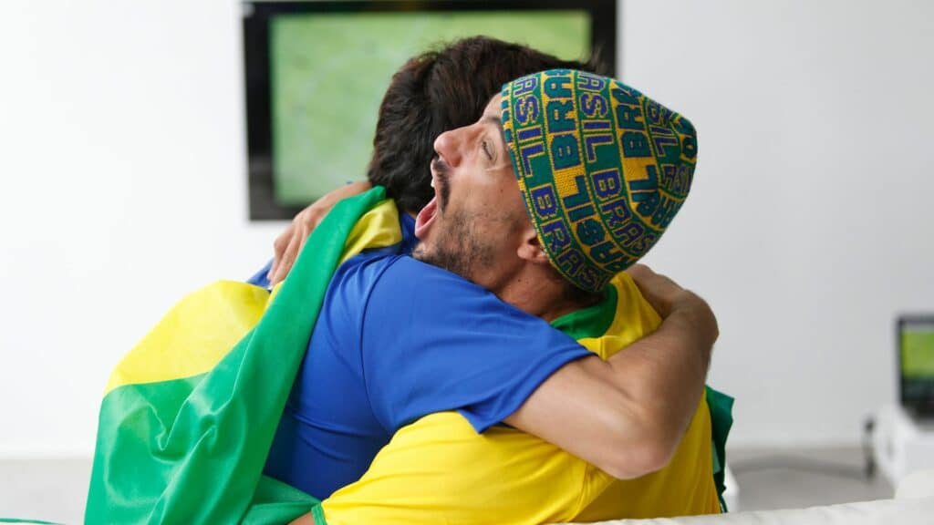Imagem de dois torcedores do Brasil, um com a bandeira nas costas e o outro com a touca do Brasil, se abraçando na frente da TV, como se comemorassem um gol.