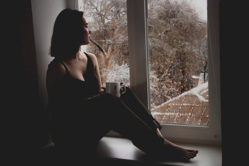 Garota sentada de frente para uma janela, pensativa, segurando uma caneca