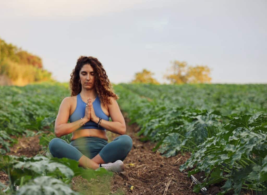 Mulher meditando sentada sobre a terra, com plantas ao seu redor.