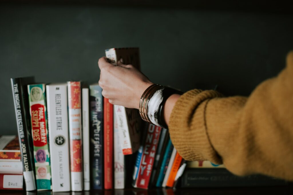 Mão feminina retirando o livro de uma prateleira com outros livros postos um ao lado do outro.