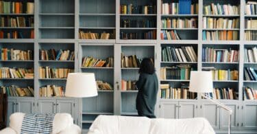 Mulher observando os livros de uma enorme estante. Há dois sofás brancos um pouco à frente da estante.