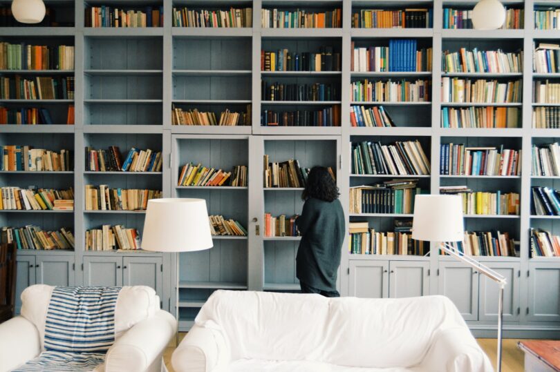 Mulher observando os livros de uma enorme estante. Há dois sofás brancos um pouco à frente da estante.