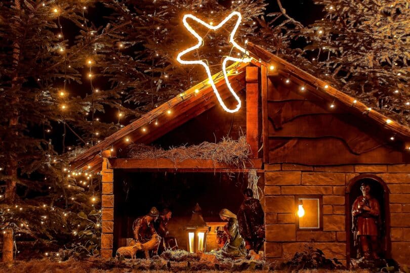 Imagem de um presépio em uma casa de madeira pequena e pisca-pisca em volta, uma estrela brilhante, feita de pisca-pisca em cima da casa.