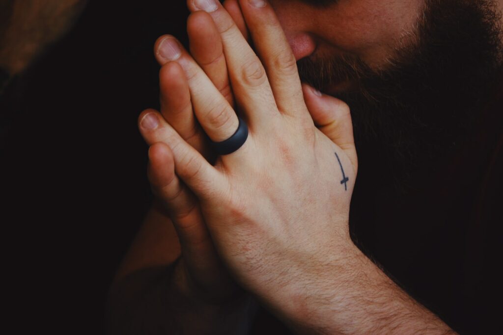 Mãos de um homem em posição de reza. Em um dos dorsos, há uma cruz tatuada.