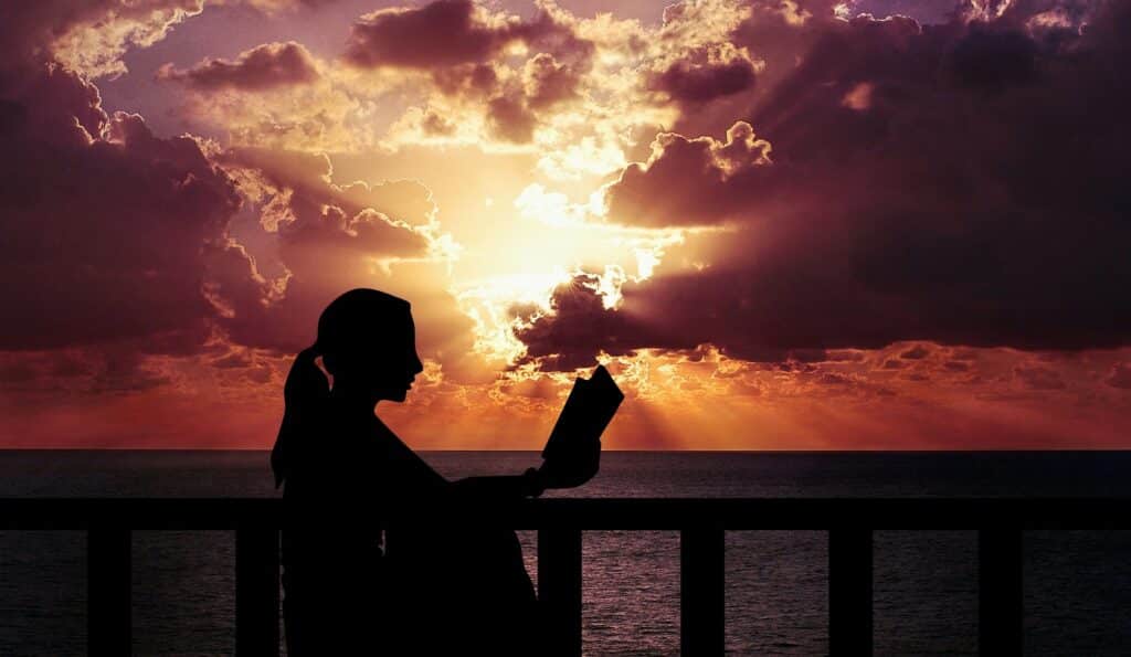Silhueta de mulher lendo um livro sob um pôr do sol na frente do mar.