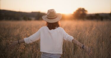 Mulher de costas em campo na natureza, de braços abertos, no pôr do sol